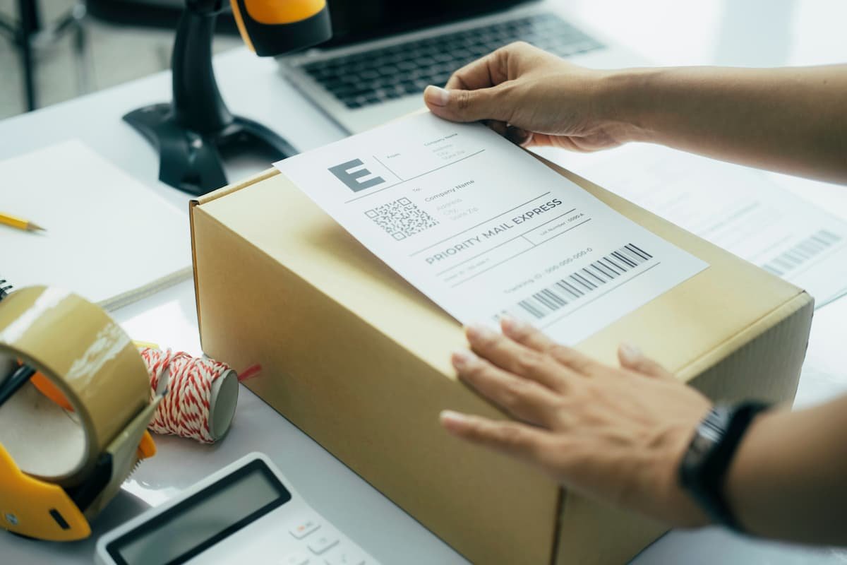 Hands placing a shipping label on a cardboard parcel on a desk with packing tape, string, calculator, laptop and barcode scanner.