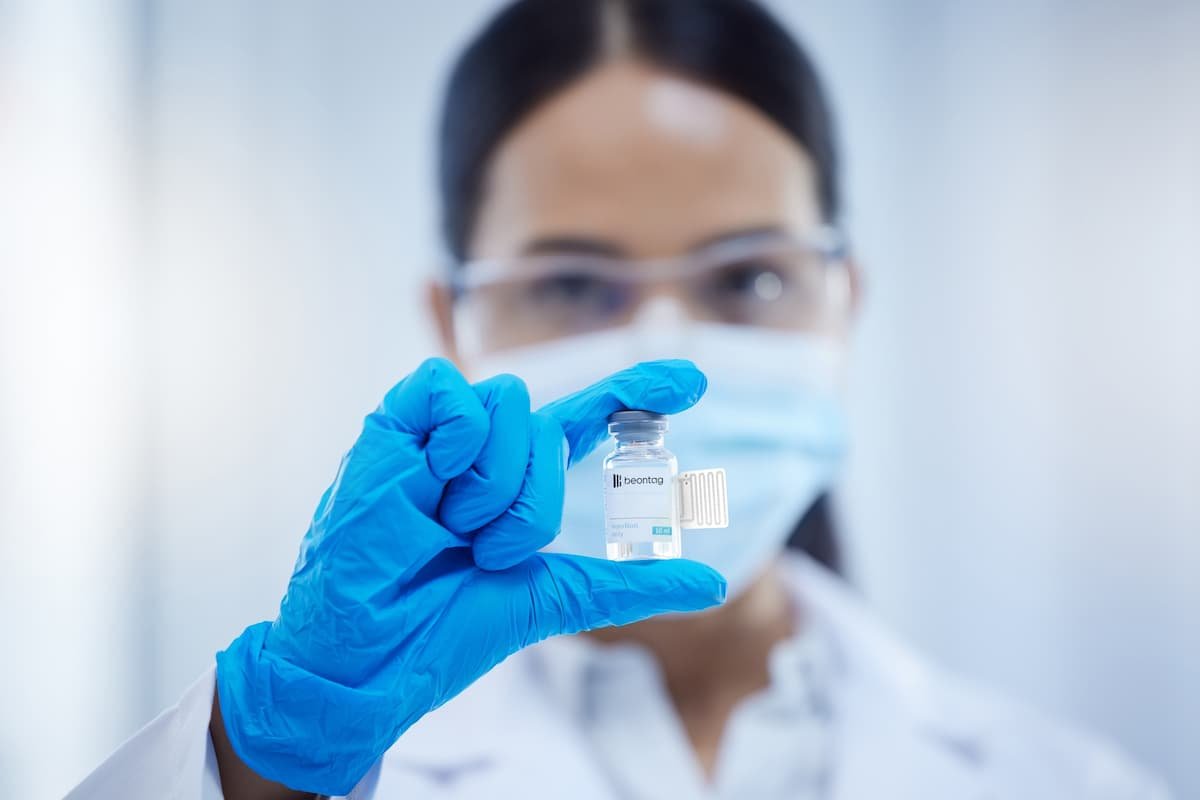 Lab technician wearing a mask and gloves holding a small vial with a Beontag label and attached RFID tag in a clinical setting.