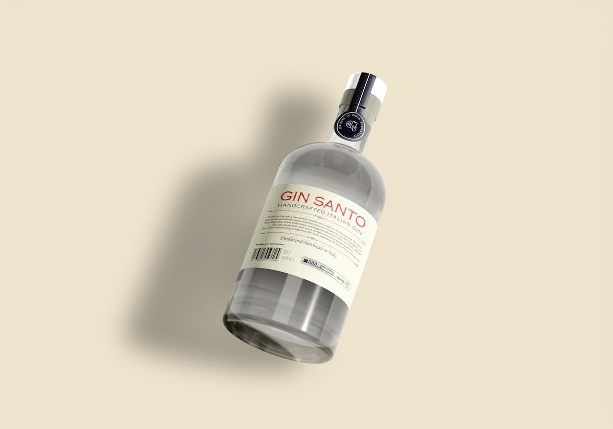 Garrafa de gin transparente, rotulada como “Gin Santo – Handcrafted Italian Gin”, exibida inclinada sobre fundo bege claro, com destaque para o rótulo de segurança de bebidas aplicado na embalagem.
