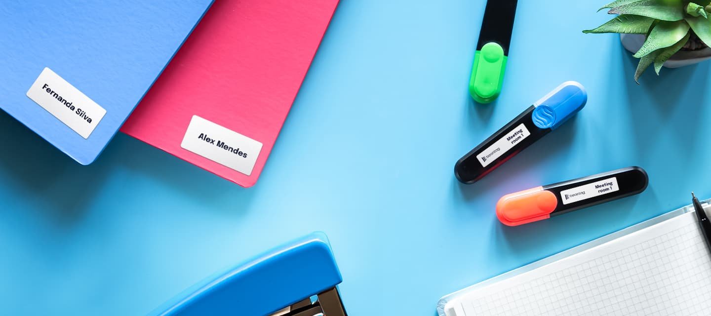 Flat lay of two colored folders with printed name labels, three handheld label maker pens, a notebook, and a succulent on a blue surface.