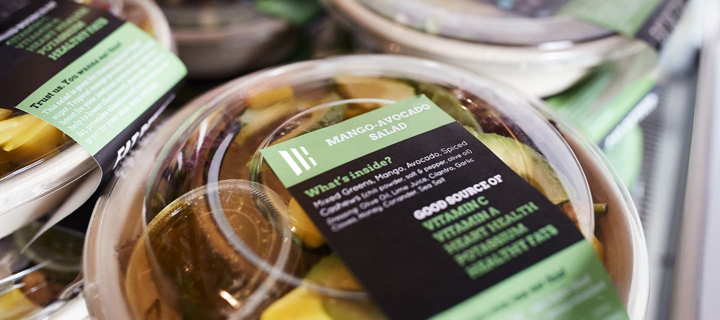 Embalagens de salada pronta em potes plásticos com tampa transparente e rótulo “Mango-Avocado Salad”, destacando o uso de adesivo hot melt na rotulagem, com outras unidades ao fundo.